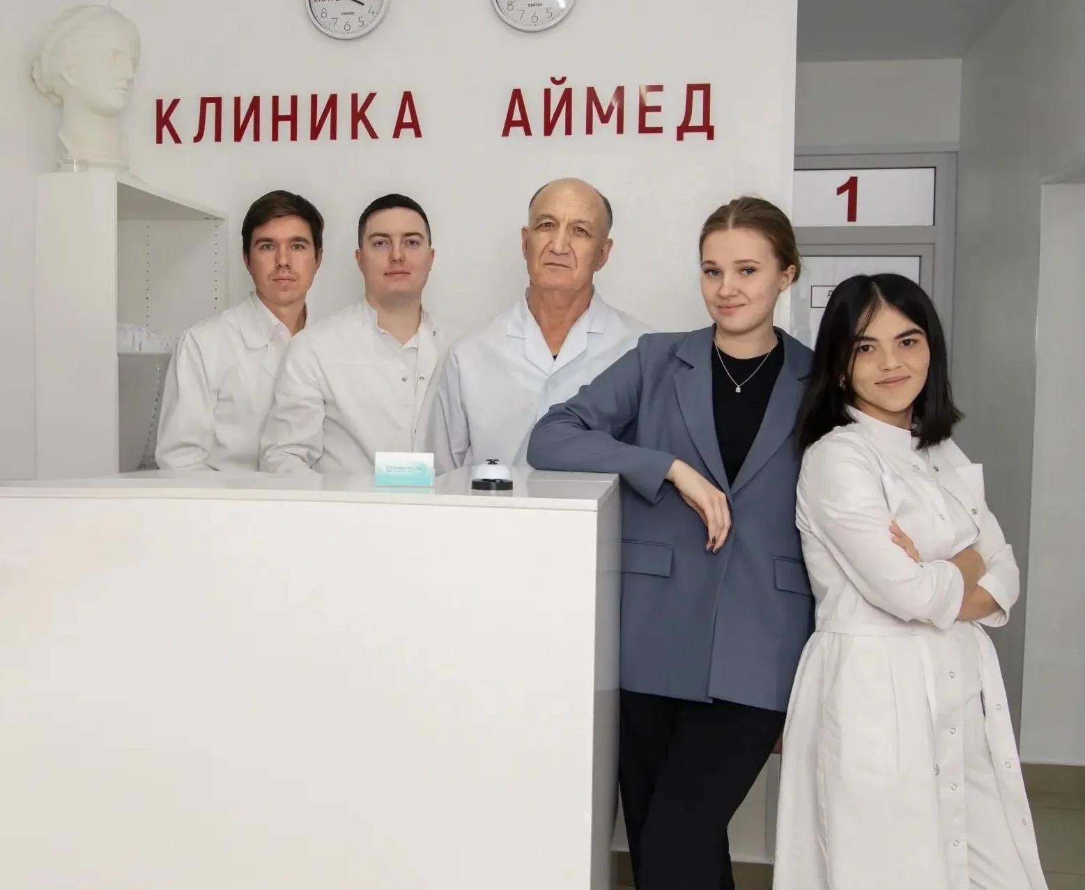 Группа медицинских специалистов, позирующих в клинике. Они одеты в белые халаты или медицинскую форму. В группе четыре женщины и трое мужчин, один человек сидит.